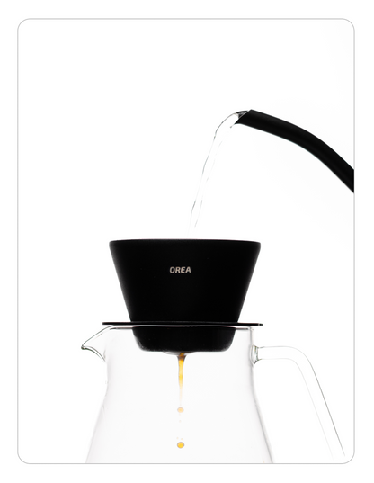 Water being poured from a thin-necked kettle into the Orea brewer, with fresh coffee dripping into a glass carafe below.