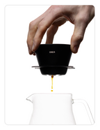 A person holding the Orea brewer by its top rim with their thumb and fingers, showing the coffee extraction process in mid-air above a carafe.