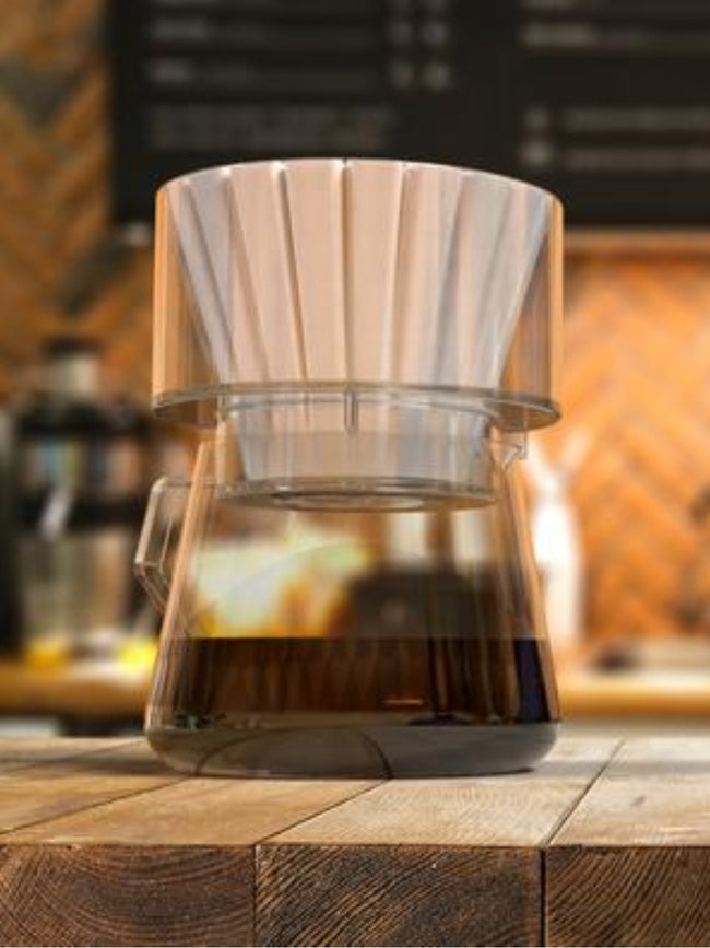 A Bathtub Simplify Brewer containing a white fluted filter, sitting atop a glass carafe filled with brewed coffee on a wooden surface.