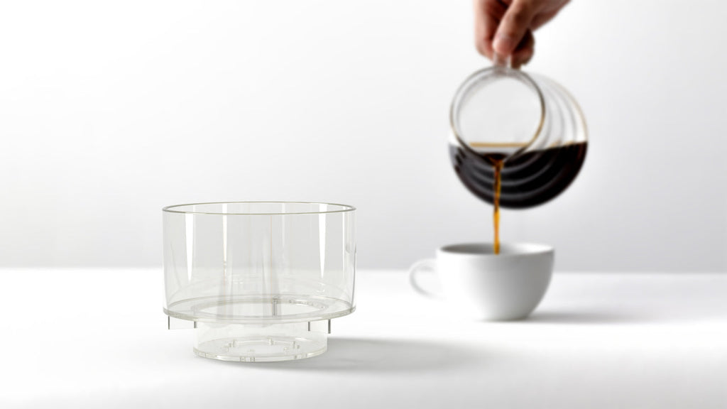 A person pouring freshly brewed coffee from a glass carafe into a white ceramic cup, with the empty Bathtub Simplify Brewer visible in the foreground.