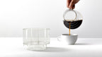 A person pouring freshly brewed coffee from a glass carafe into a white ceramic cup, with the empty Bathtub Simplify Brewer visible in the foreground.