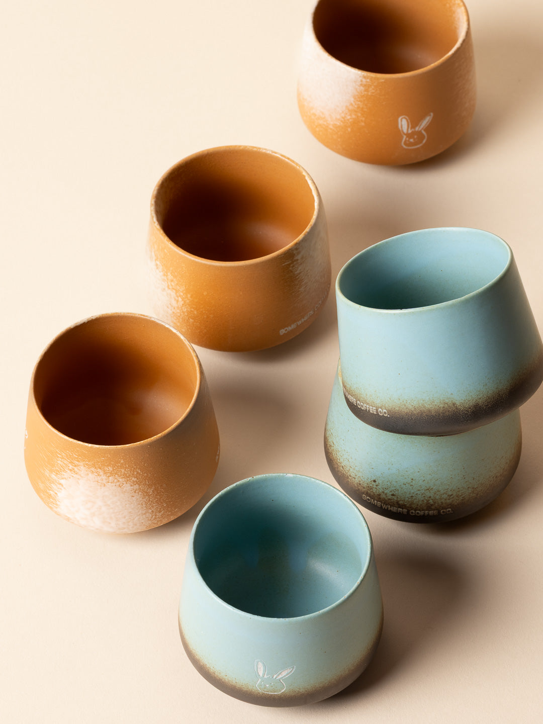 A creative overhead arrangement of Somewhere Coffee Co.'s 200ml artisan ceramic cups, showcasing the tactile handleless design and the rich, reactive glaze variations across the teal and orange collections.