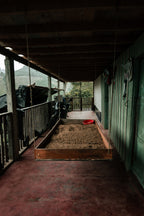 A rustic wooden raised drying bed filled with drying parchment coffee sits on the farmhouse porch at La Cabaña Farm in Bruselas, Huila, flanked by a parked motorcycle and hanging ropes, showcasing the artisanal, farm-direct nature of Somewhere Coffee's sourcing.