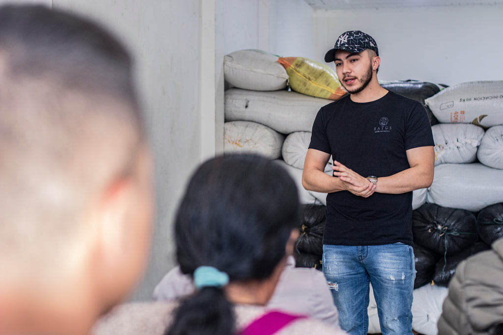 Bryan Smith, wearing a black t-shirt and baseball cap, standing in a warehouse filled with stacked white coffee bags, passionately addressing an audience, highlighting the direct-trade transparency and community-driven approach of Finca Satus for Somewhere Coffee.