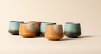 A beautiful lineup of five handleless ceramic coffee cups from Somewhere Coffee Co., alternating between the teal and amber reactive glaze finishes, each featuring the signature hand-drawn rabbit motif and matte texture.