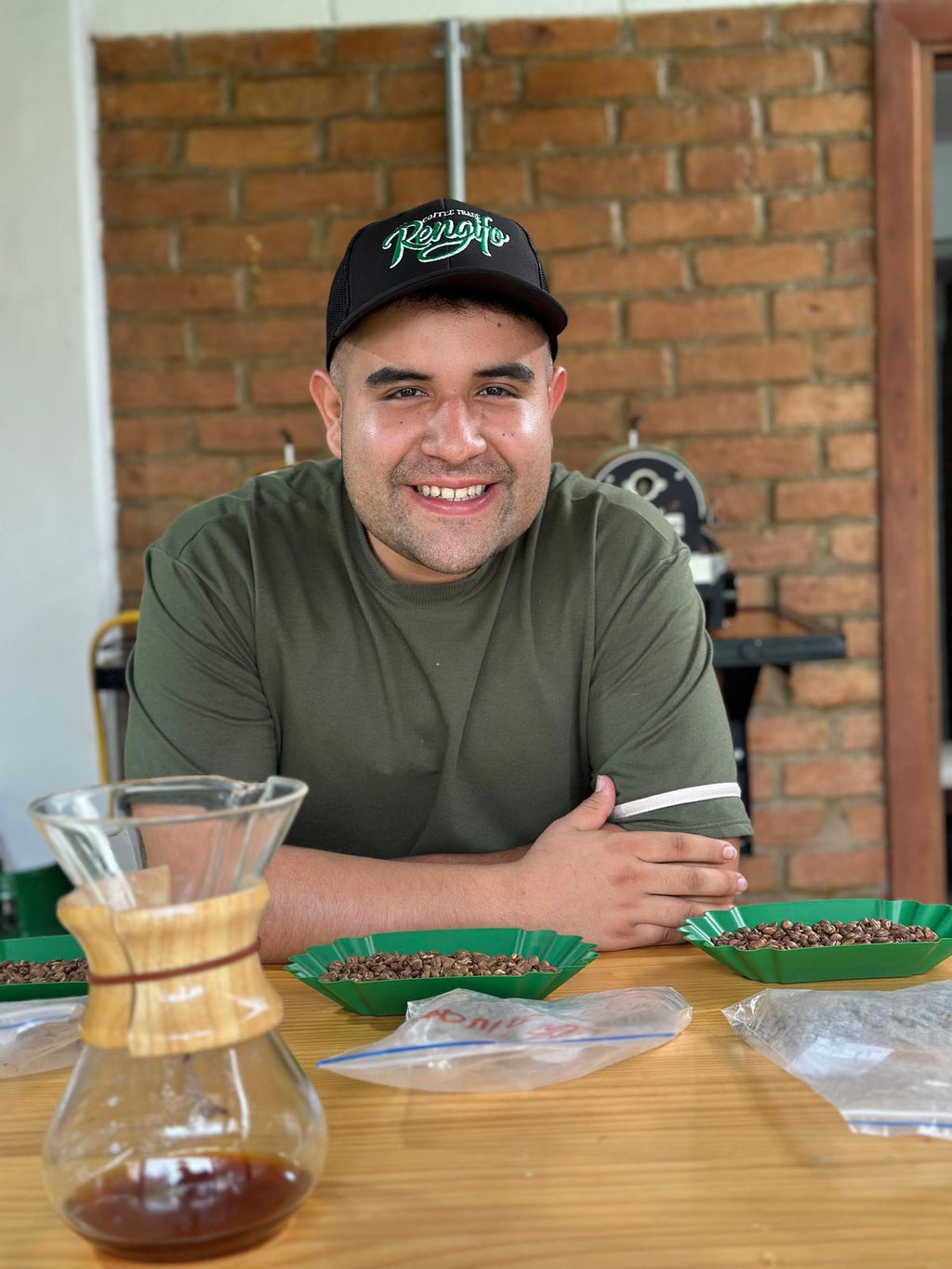 A coffee producer smiling at the cupping table, presenting freshly brewed Gesha coffee in a glass Chemex at Laboyani Farm in Pitalito, Huila, Colombia, for Somewhere Coffee.