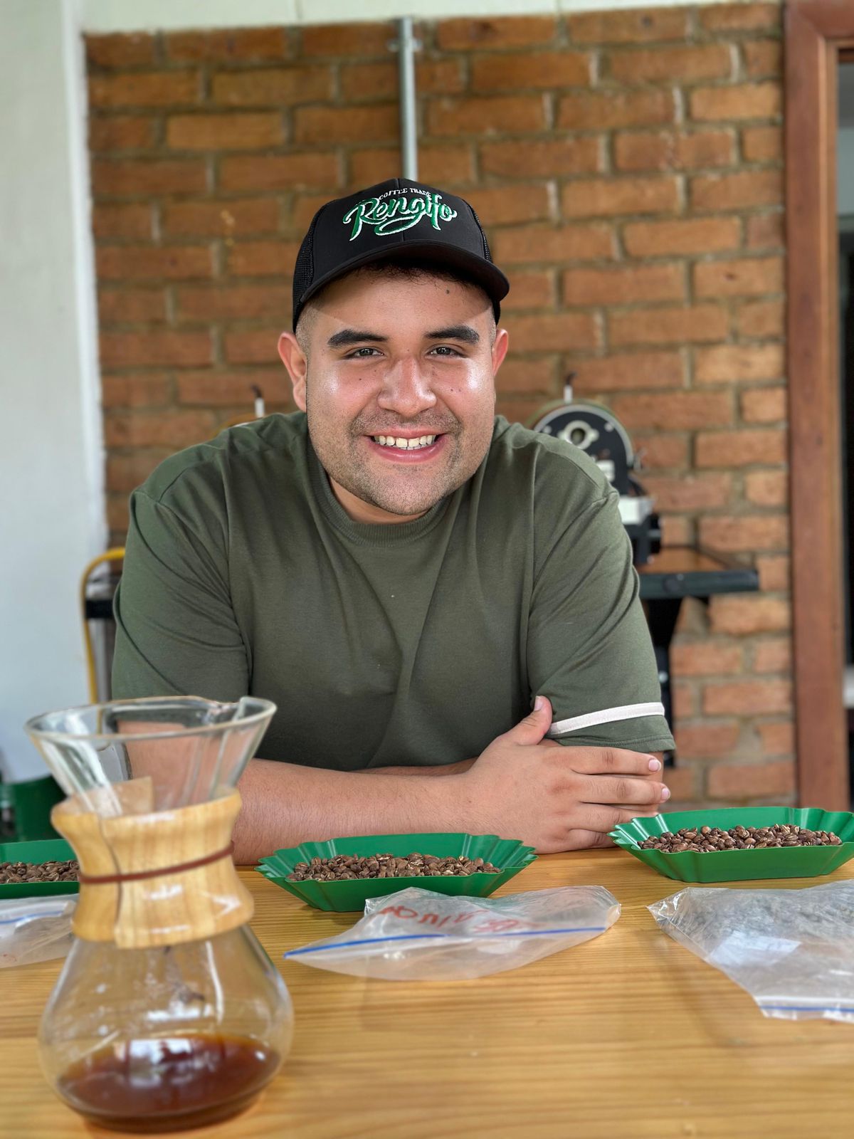 A coffee producer smiling at the cupping table, presenting freshly brewed Gesha coffee in a glass Chemex at Laboyani Farm in Pitalito, Huila, Colombia, for Somewhere Coffee.