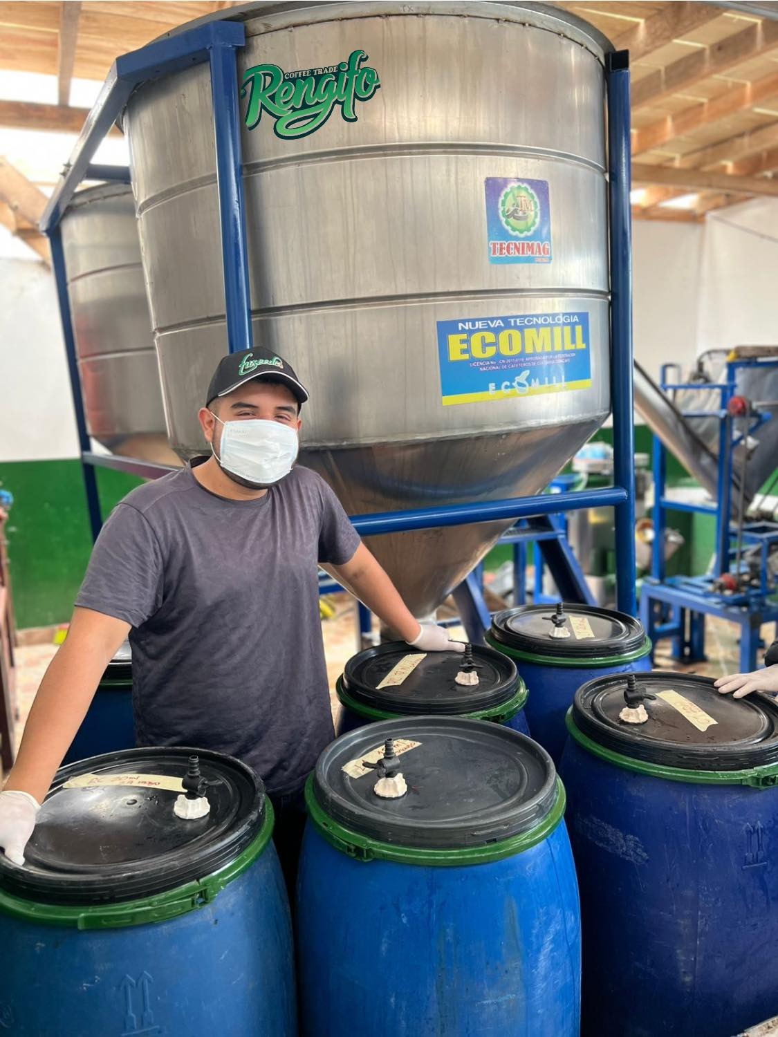 A worker wearing a mask and standing proudly beside industrial stainless steel EcoMill fermentation tanks and blue drums, showcasing the modern washed processing setup at Laboyani Farm in Pitalito, Huila.