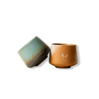 A clean, high-contrast product shot of two handleless ceramic tumblers—one teal and one amber—displayed on a pure white background, highlighting the tactile design, premium reactive glazes, and artisan craftsmanship of Somewhere Coffee Co.'s pottery.