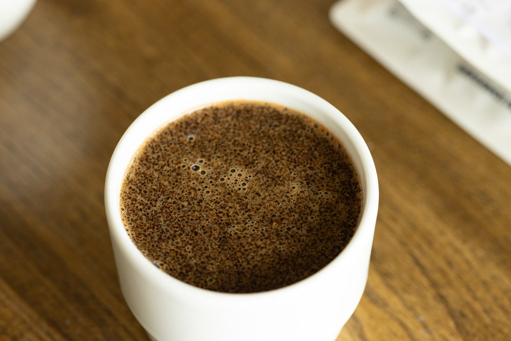 An overhead macro shot of rich, frothy coffee bloom with tiny bubbles resting inside a pristine white KAT CUP porcelain bowl, highlighting the wide, accessible opening perfect for slurping and evaluating aroma.