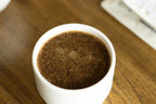 An overhead macro shot of rich, frothy coffee bloom with tiny bubbles resting inside a pristine white KAT CUP porcelain bowl, highlighting the wide, accessible opening perfect for slurping and evaluating aroma.