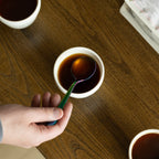 A hand holding a vibrant, iridescent rainbow-colored spoon over a white KAT CUP filled with dark coffee, showcasing a modern, colorful twist on the traditional specialty coffee cupping experience.