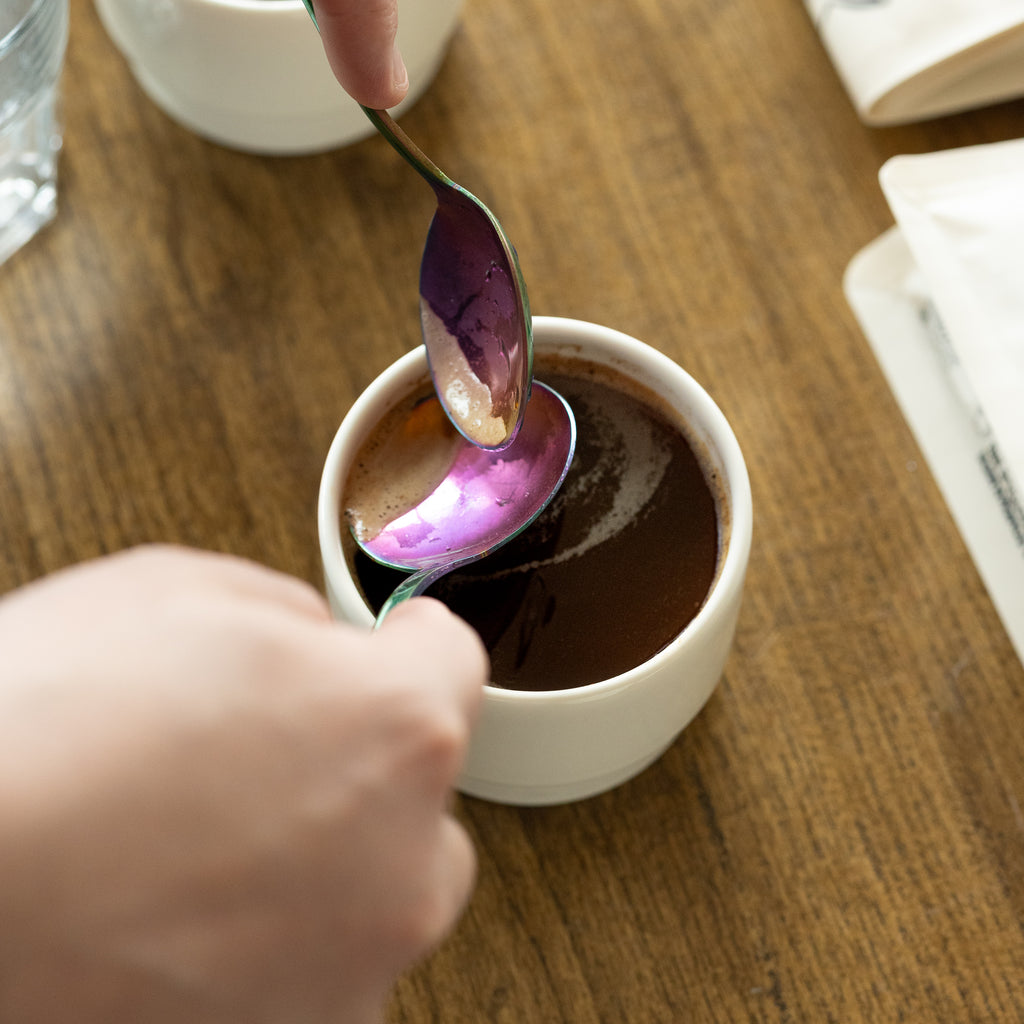 A close-up action shot of an iridescent spoon breaking the surface of freshly brewed coffee inside a white Porland KAT CUP, capturing the sensory ritual of tasting and evaluating coffee.