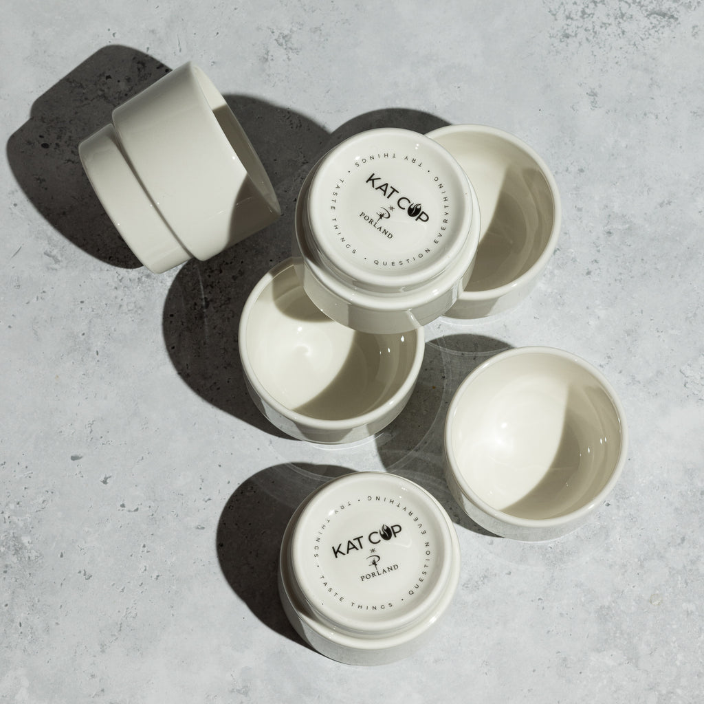 An artistic overhead flat lay of several white Porland KAT CUPs, showing them both upright and inverted on a textured concrete surface, highlighting the stark white porcelain against the grey background.