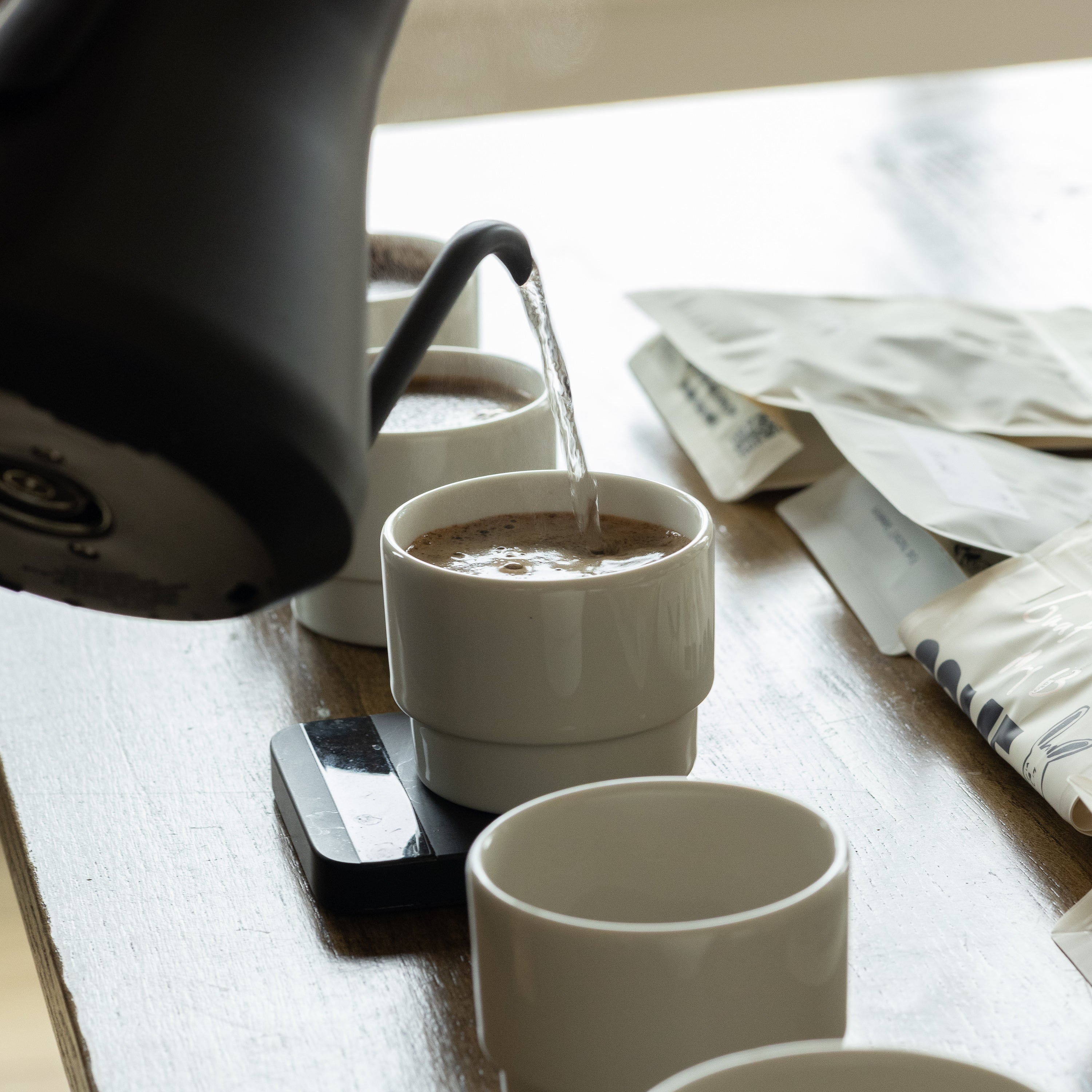 A close-up of a black gooseneck kettle pouring hot water into a white Porland KAT CUP resting on a digital scale, demonstrating its use for precise manual pour-over brewing with Somewhere Coffee beans.