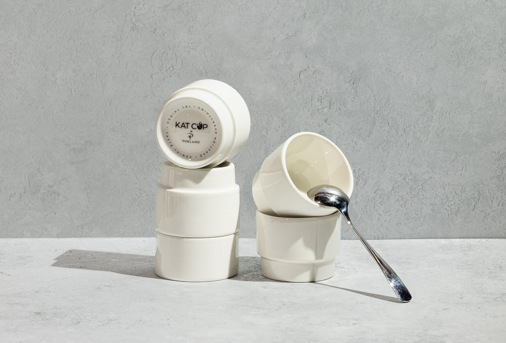 A beautifully stylized still-life of stacked white Porland KAT CUPs on a concrete surface, with one cup leaning down revealing its bottom stamp, and a sleek silver spoon resting elegantly inside the top cup.