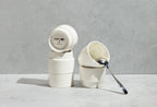 A beautifully stylized still-life of stacked white Porland KAT CUPs on a concrete surface, with one cup leaning down revealing its bottom stamp, and a sleek silver spoon resting elegantly inside the top cup.
