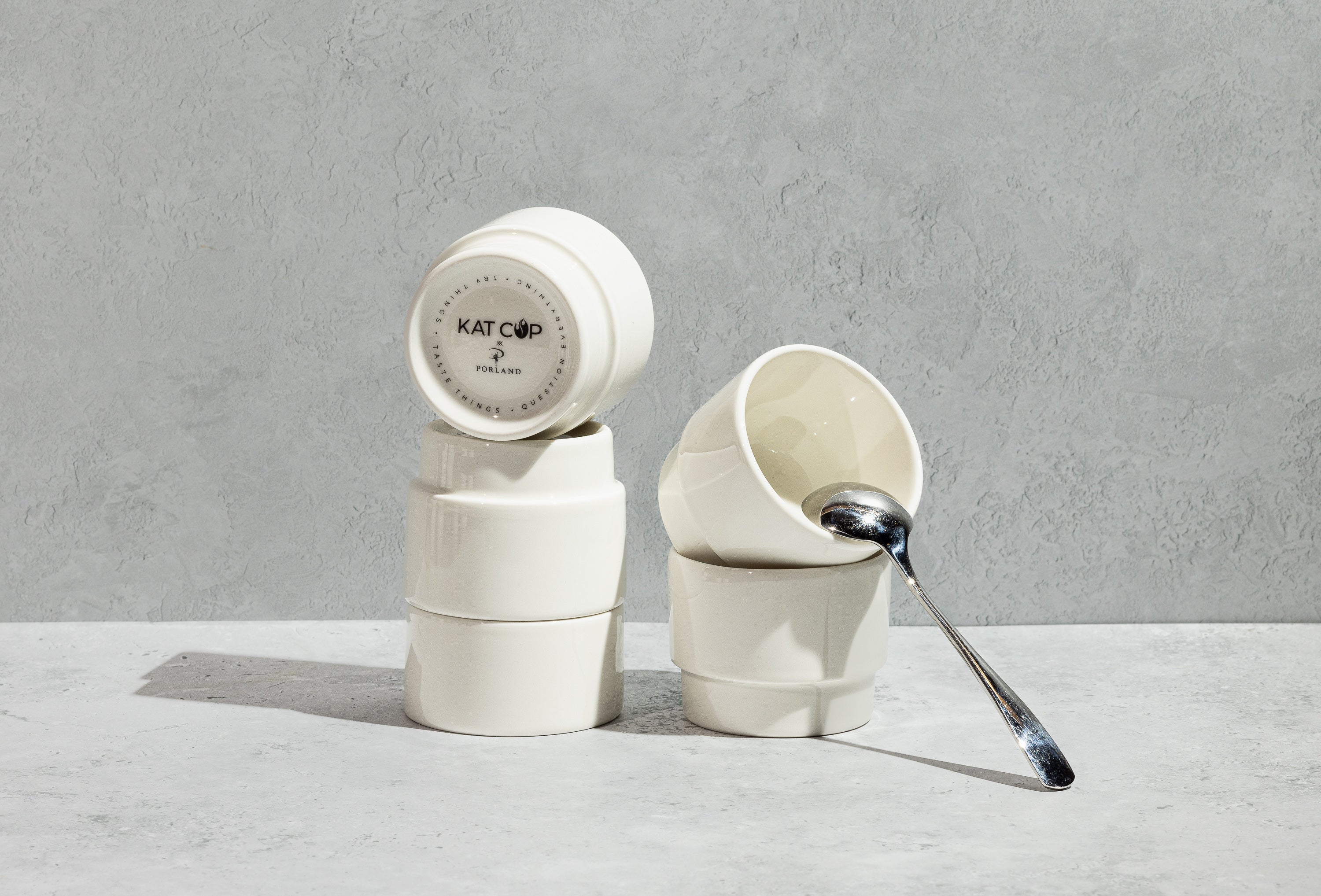 A beautifully stylized still-life of stacked white Porland KAT CUPs on a concrete surface, with one cup leaning down revealing its bottom stamp, and a sleek silver spoon resting elegantly inside the top cup.