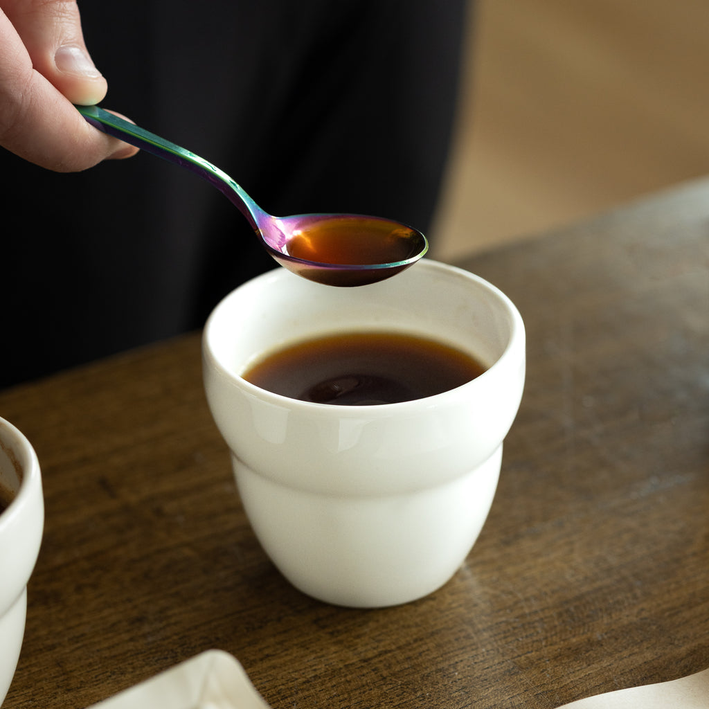 A close-up of a hand holding a vibrant, iridescent rainbow-colored spoon filled with brewed coffee above a white Roaster Kat KatCup Omni, capturing the sensory ritual of professional cupping and coffee evaluation.