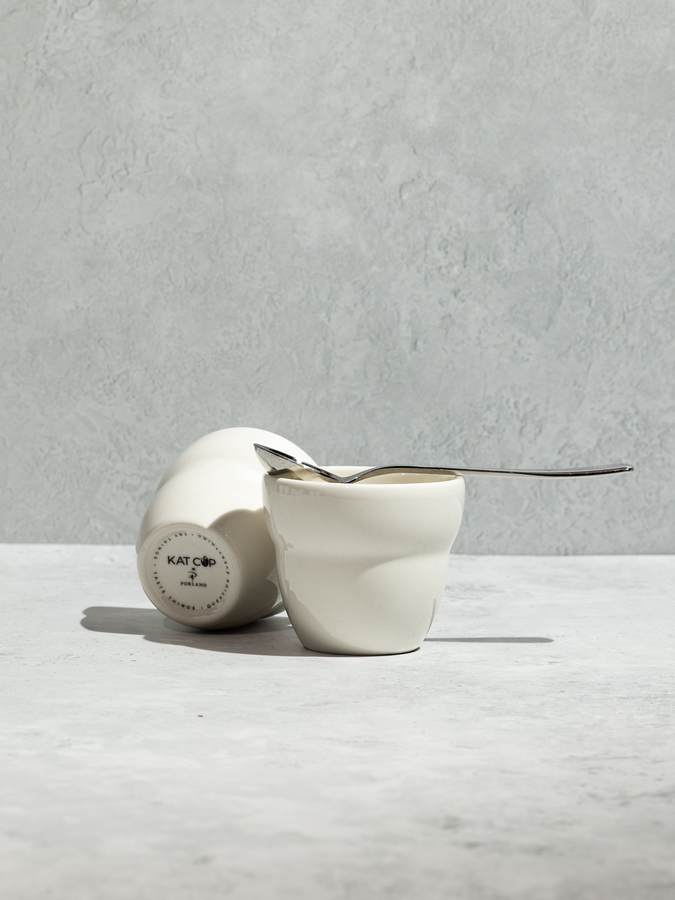A clean studio still-life of two white Roaster Kat KatCup Omni porcelain bowls on a textured concrete surface, with one resting upright holding a silver tasting spoon and the other showing the sleek stamped base.