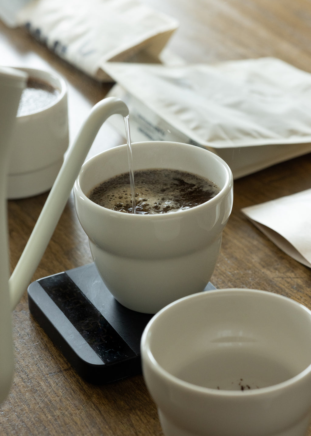 A lifestyle action shot of hot water being carefully poured from a white gooseneck kettle into a 190ml KatCup Omni resting on a black scale, demonstrating its perfect design for precision filter coffee brewing.