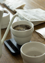 A lifestyle action shot of hot water being carefully poured from a white gooseneck kettle into a 190ml KatCup Omni resting on a black scale, demonstrating its perfect design for precision filter coffee brewing.