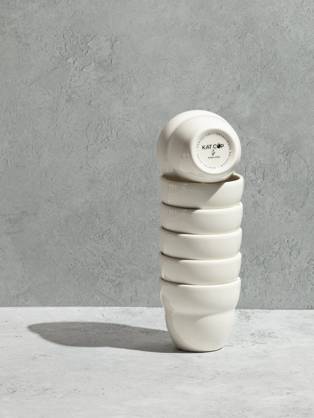 A beautifully composed studio shot showcasing the exceptional stackability of the KatCup Omni, featuring a tall, perfectly balanced tower of six white porcelain cups resting on a concrete counter, with the bottom stamp clearly visible on the top inverted cup.