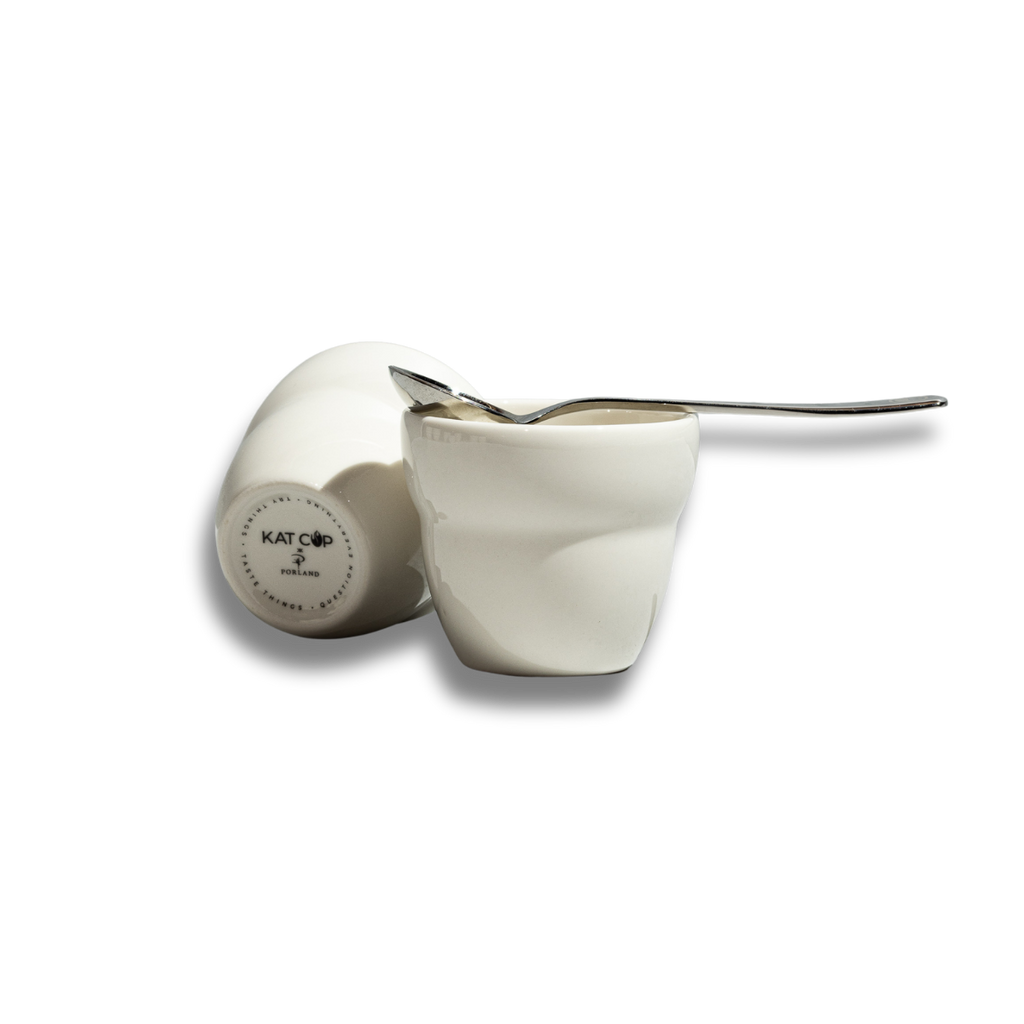A clean product cutout on a stark white background of two white Roaster Kat KatCup Omnis, featuring one upright cup holding a silver spoon and one tilted cup displaying the "KAT CUP PORLAND" circular stamp on its base.