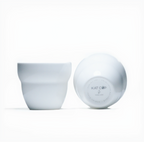 A bright, minimalist studio product shot of a white Roaster Kat KatCup Omni in two positions—upright showing its ergonomic, hand-friendly shape, and tilted on its side displaying the distinct KAT CUP base logo.