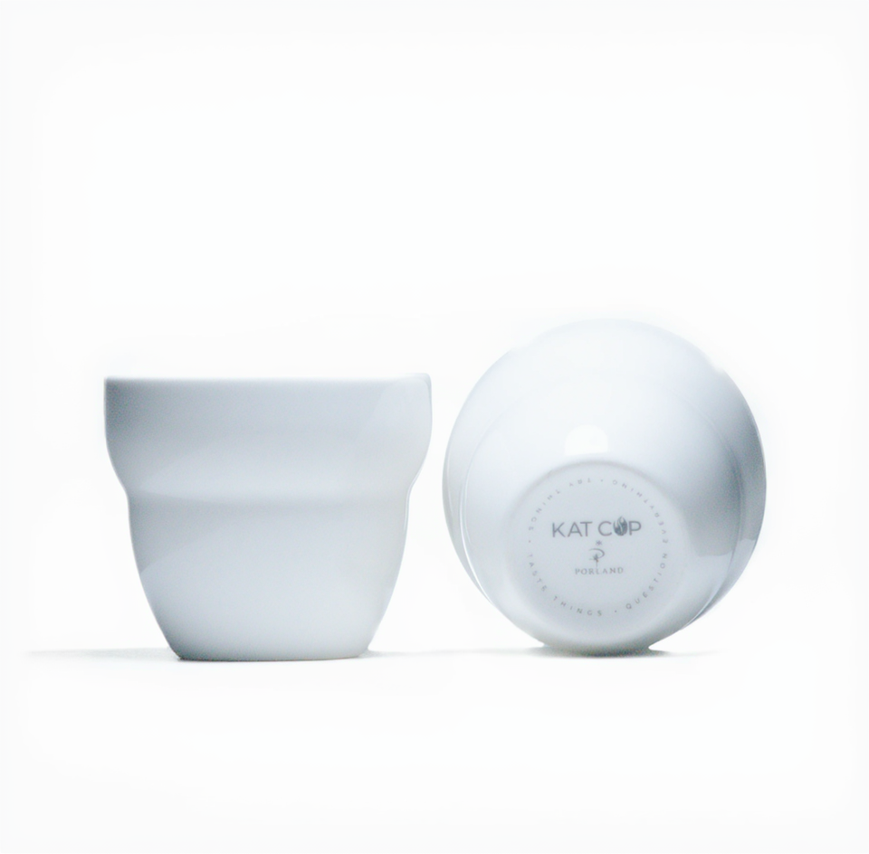 A bright, minimalist studio product shot of a white Roaster Kat KatCup Omni in two positions—upright showing its ergonomic, hand-friendly shape, and tilted on its side displaying the distinct KAT CUP base logo.