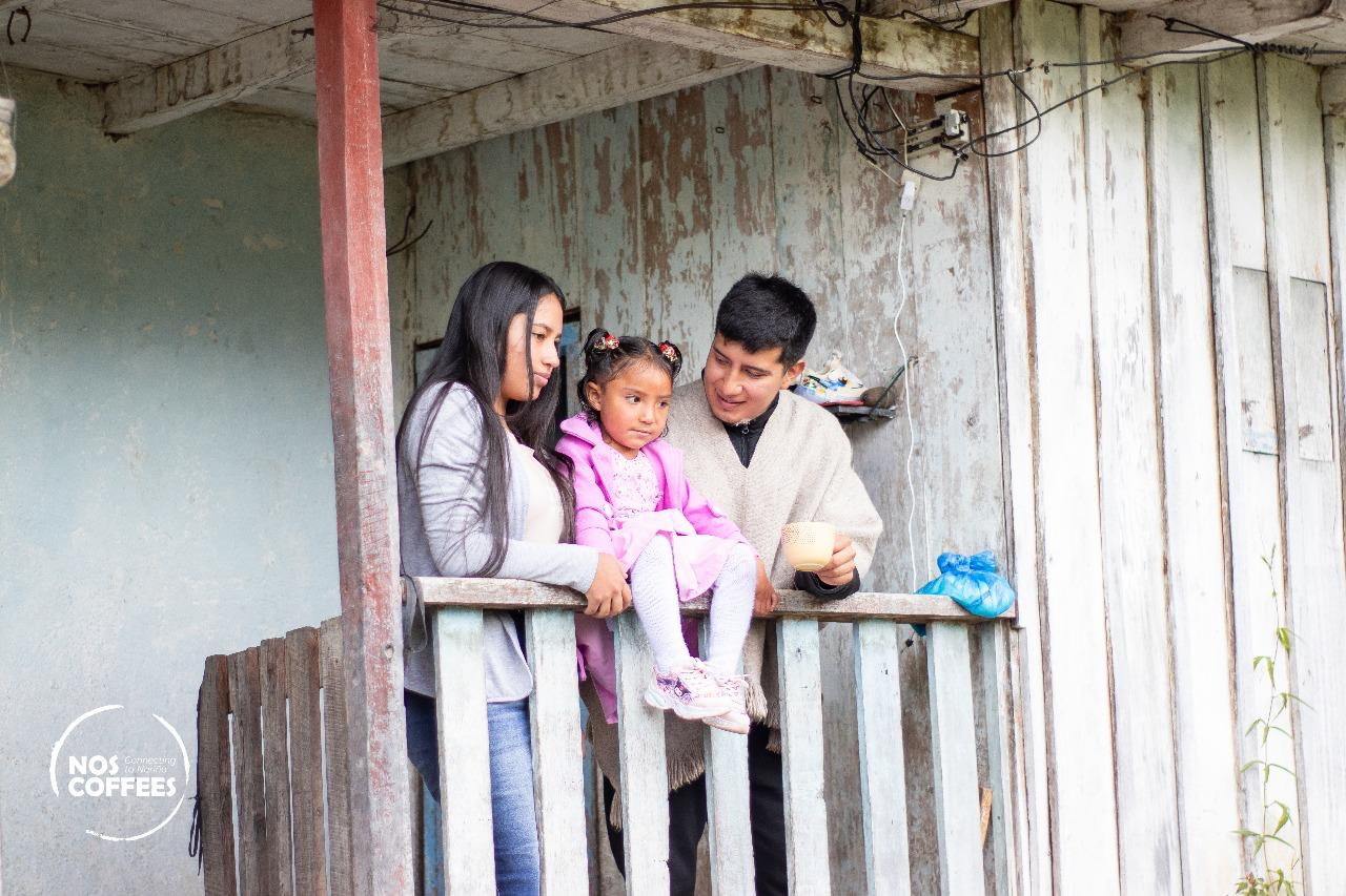 A candid moment of a young indigenous coffee-producing family standing on the rustic, weathered wooden porch of their home in Nariño, Colombia. The father, wearing a traditional wool poncho and holding a small cup of coffee, represents the generational craftsmanship and community behind Somewhere Coffee's high-altitude washed Castillo lot.