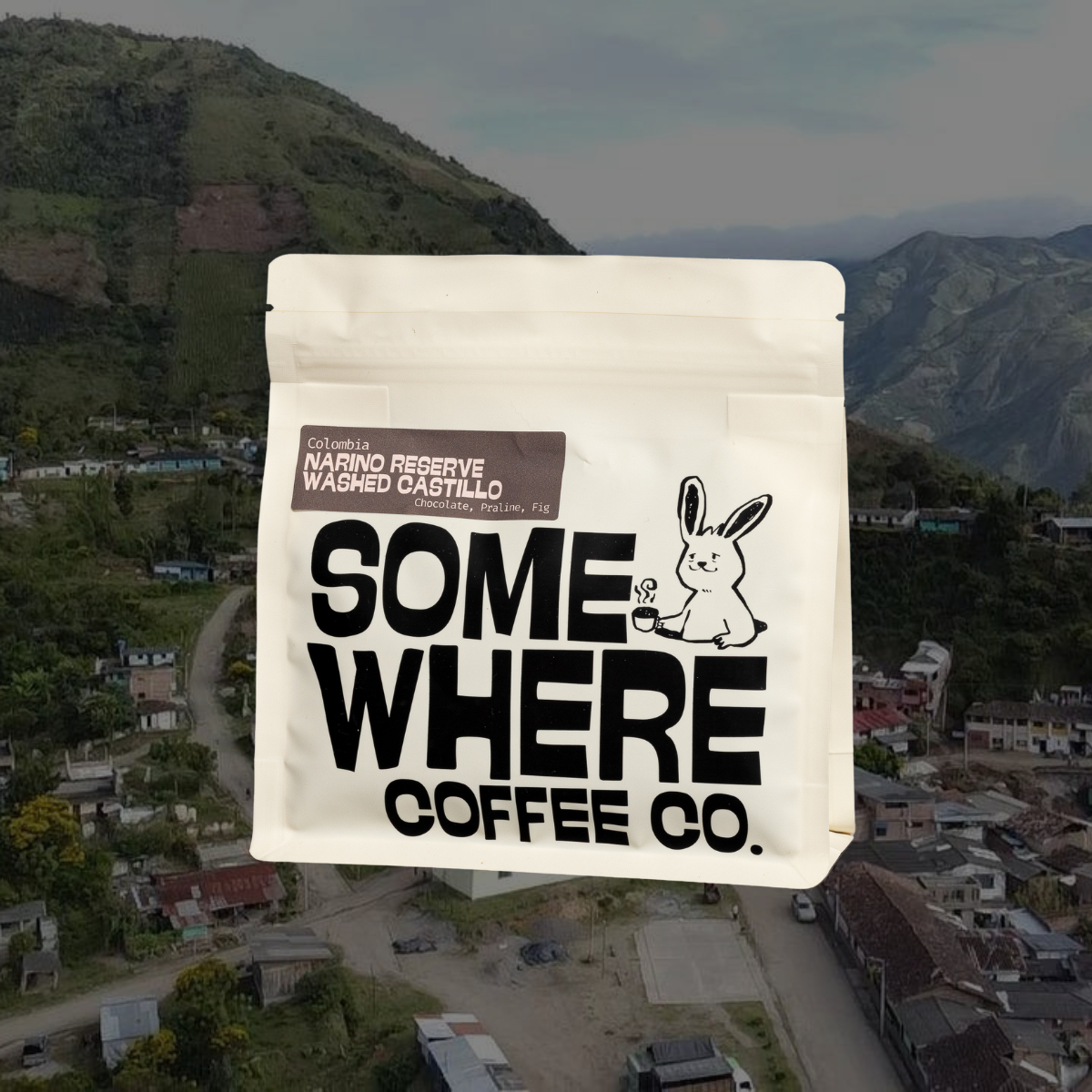 A clean product shot of Somewhere Coffee Co.'s white signature bag, featuring the rabbit logo, placed against a breathtaking backdrop of the Nariño mountain landscape. The bag's label reads "Nariño Reserve Washed Castillo" with tasting notes of Chocolate, Praline, and Fig.