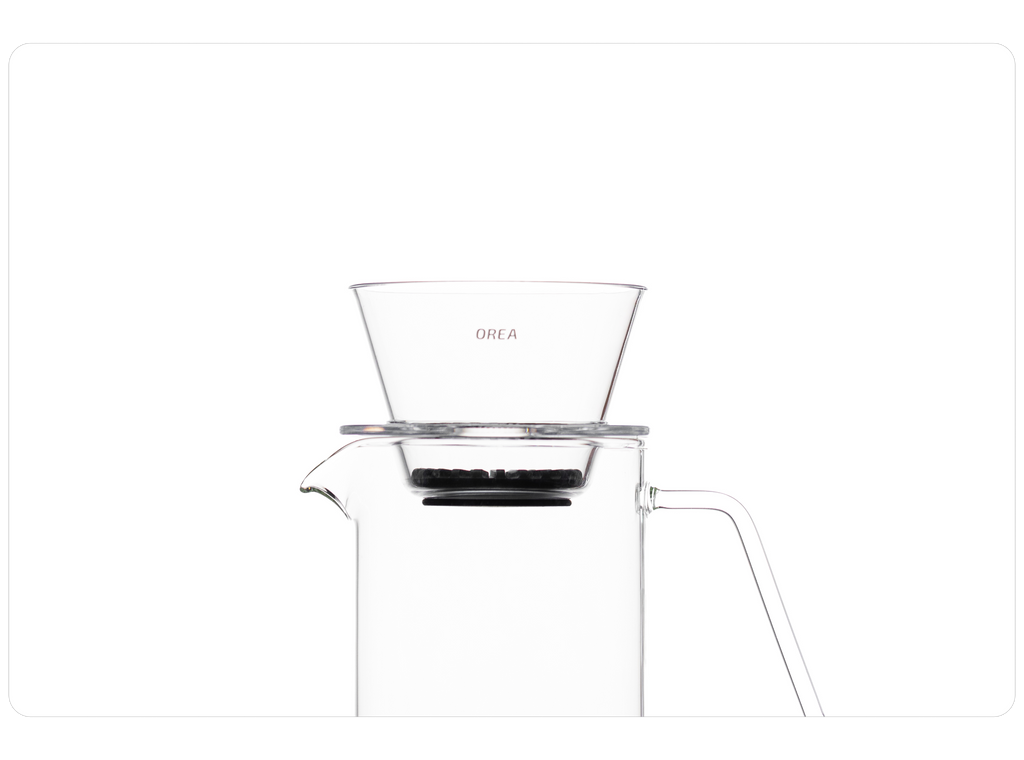A clean studio shot of a clear OREA O2 brewer resting directly on the rim of a clear glass pour-over carafe, showcasing its flat-bottom design and universal fit for a seamless brewing setup.