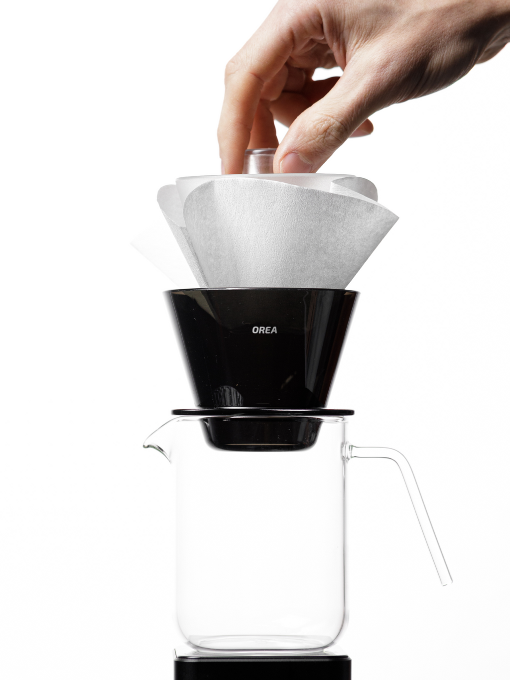 A hand pressing down to pre-wet and seat a large 185 wave paper filter into the OREA O2 Brewer, demonstrating how the open bottom allows the paper to flex downward and properly seal against the brewer walls.