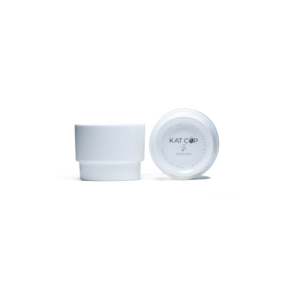 A clean, bright studio product shot of a white Porland KAT CUP in two views—one upright showing its minimalist cylindrical shape, and one inverted displaying the distinctive KAT CUP logo and stamp on the base.