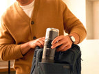 A man wearing a mustard cardigan sliding the compact OXO Brew Rapid Brewer into the side pocket of a navy blue backpack, proving it is the ultimate travel-friendly brewing tool for coffee lovers on the go.