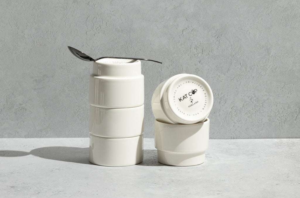 A tall, balanced stack of three white Porland KAT CUPs resting on a concrete counter, with a silver spoon resting inside the rim of the top cup, emphasizing their clever, nesting stackability for coffee cupping.
