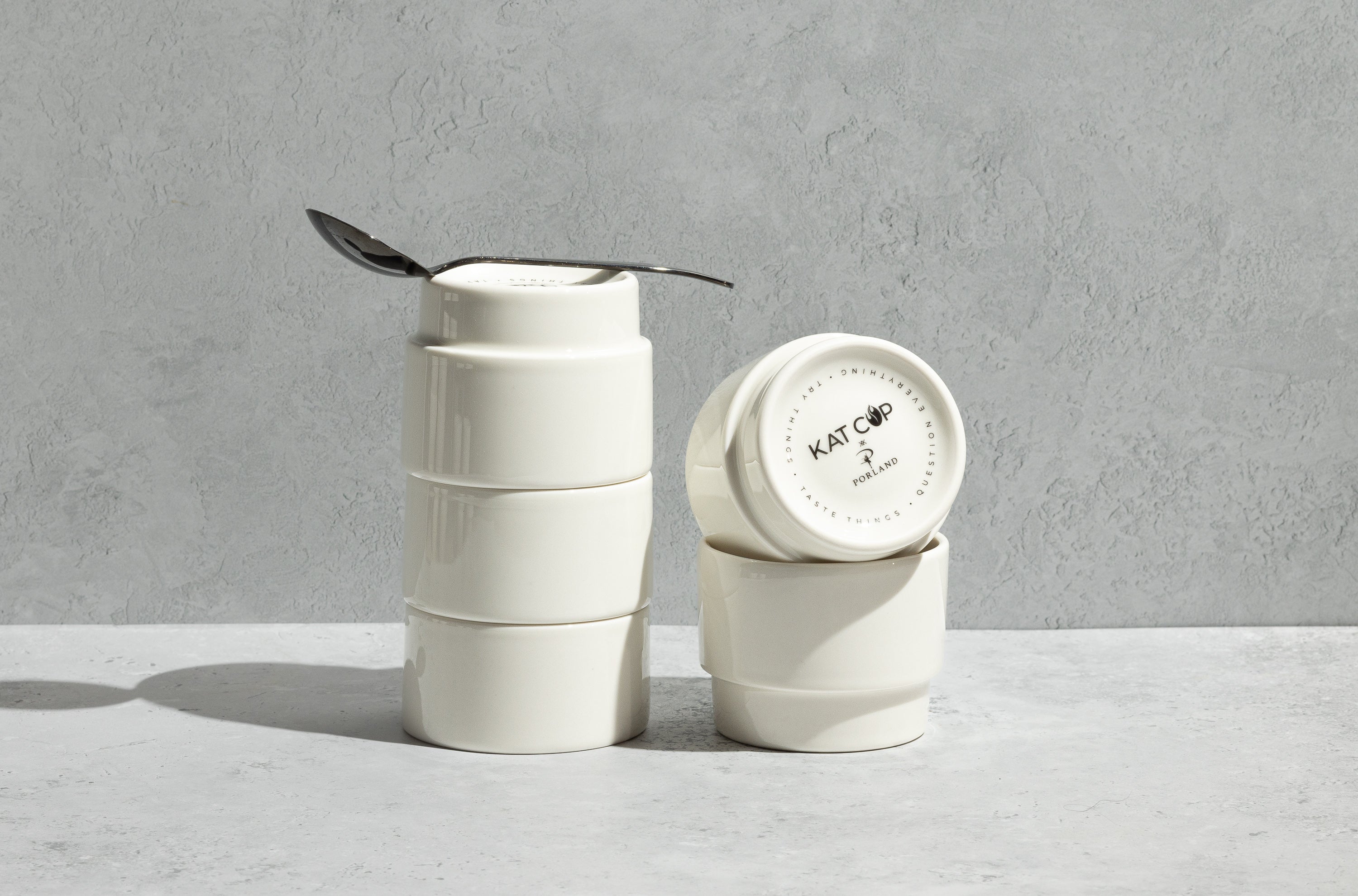 A tall, balanced stack of three white Porland KAT CUPs resting on a concrete counter, with a silver spoon resting inside the rim of the top cup, emphasizing their clever, nesting stackability for coffee cupping.