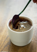 A glossy, close-up macro shot of an iridescent spoon stirring a swirling pool of dark brewed coffee inside a white KAT CUP by Porland, showcasing the cup's smooth, high-quality interior glaze.