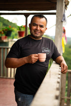 A warm, portrait-style shot of a smiling Willian Ortiz, producer of La Cabaña Farm in Bruselas, Huila, holding a ceramic coffee cup while leaning on the wooden porch railing, symbolizing the pride and craftsmanship behind Somewhere Coffee's experimental Castillo lot.