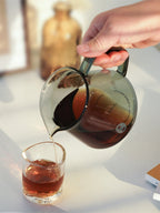 A hand pouring a clean, steady stream of freshly brewed coffee from the TIMEMORE glass server into a small drinking glass, demonstrating the smooth, precise spout control.