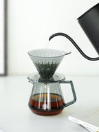 The TIMEMORE Coffee Server in a transparent black finish being brewed into with a black gooseneck kettle, showcasing a sleek, modern aesthetic for pour-over coffee.
